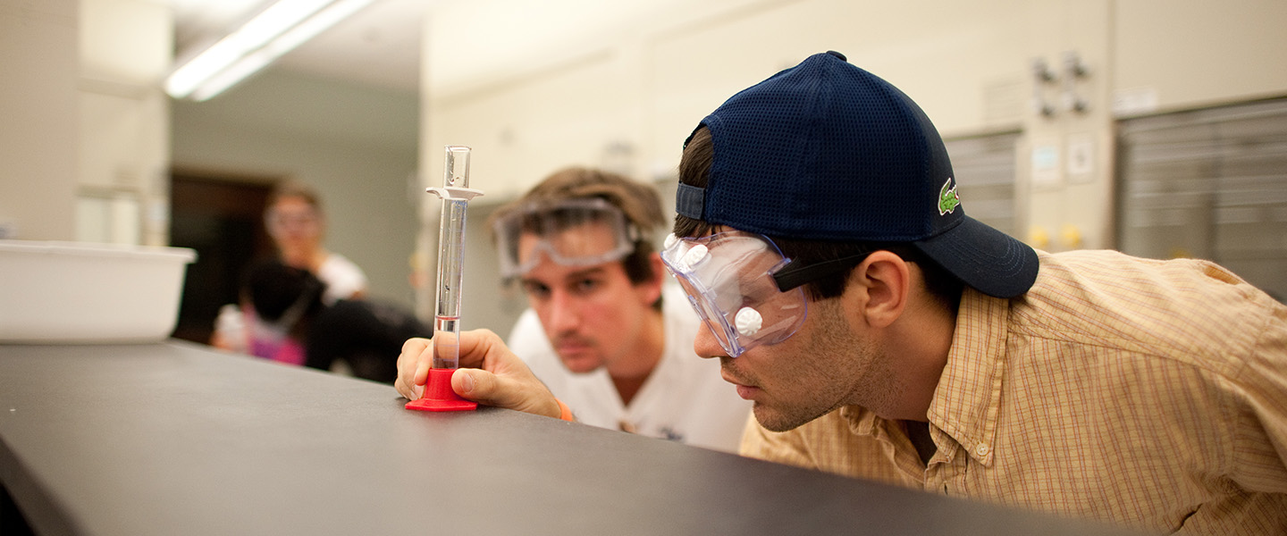 Two male students in safety goggles look at a test tube from the edge of a table.