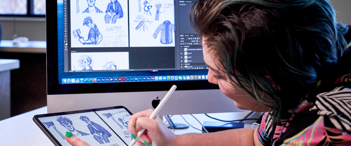Student draws illustrations on tablet next to laptop with designs on it