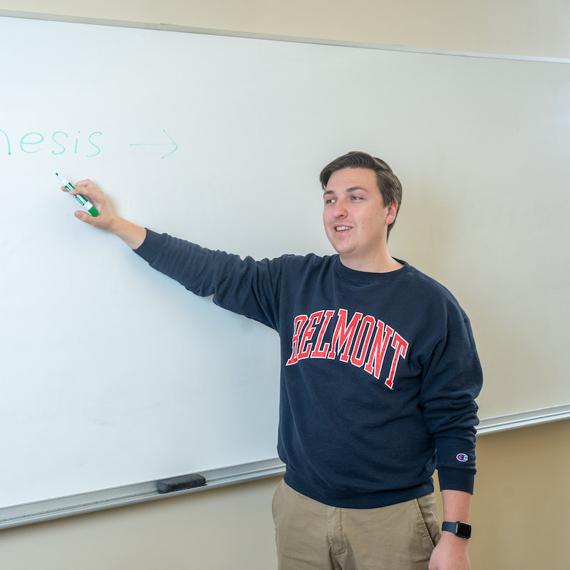 A male student in a Belmont sweatshirt stands at a whiteboard and gestures to it while speaking.