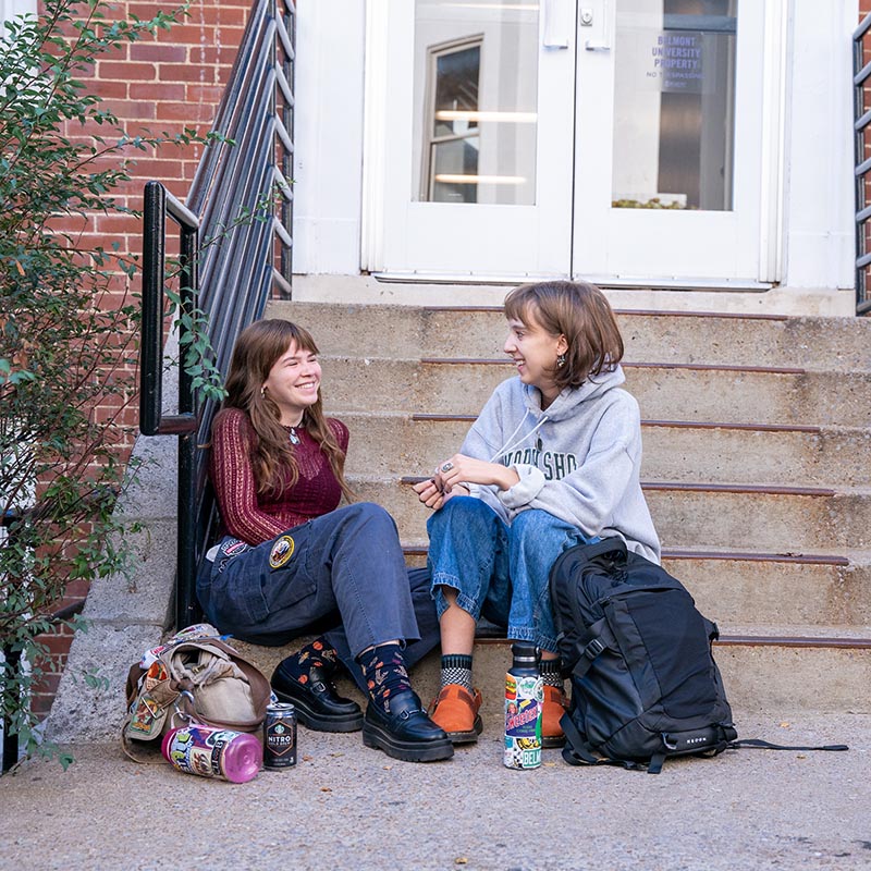 2 students sitting and talking on a concrete staircase outside of a dorm