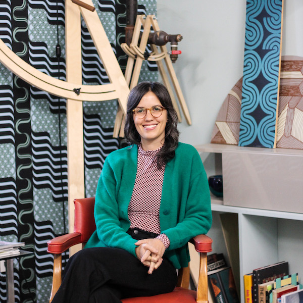 Alumna Elizabeth Williams sits in red chair in front of the colorful background of her studio