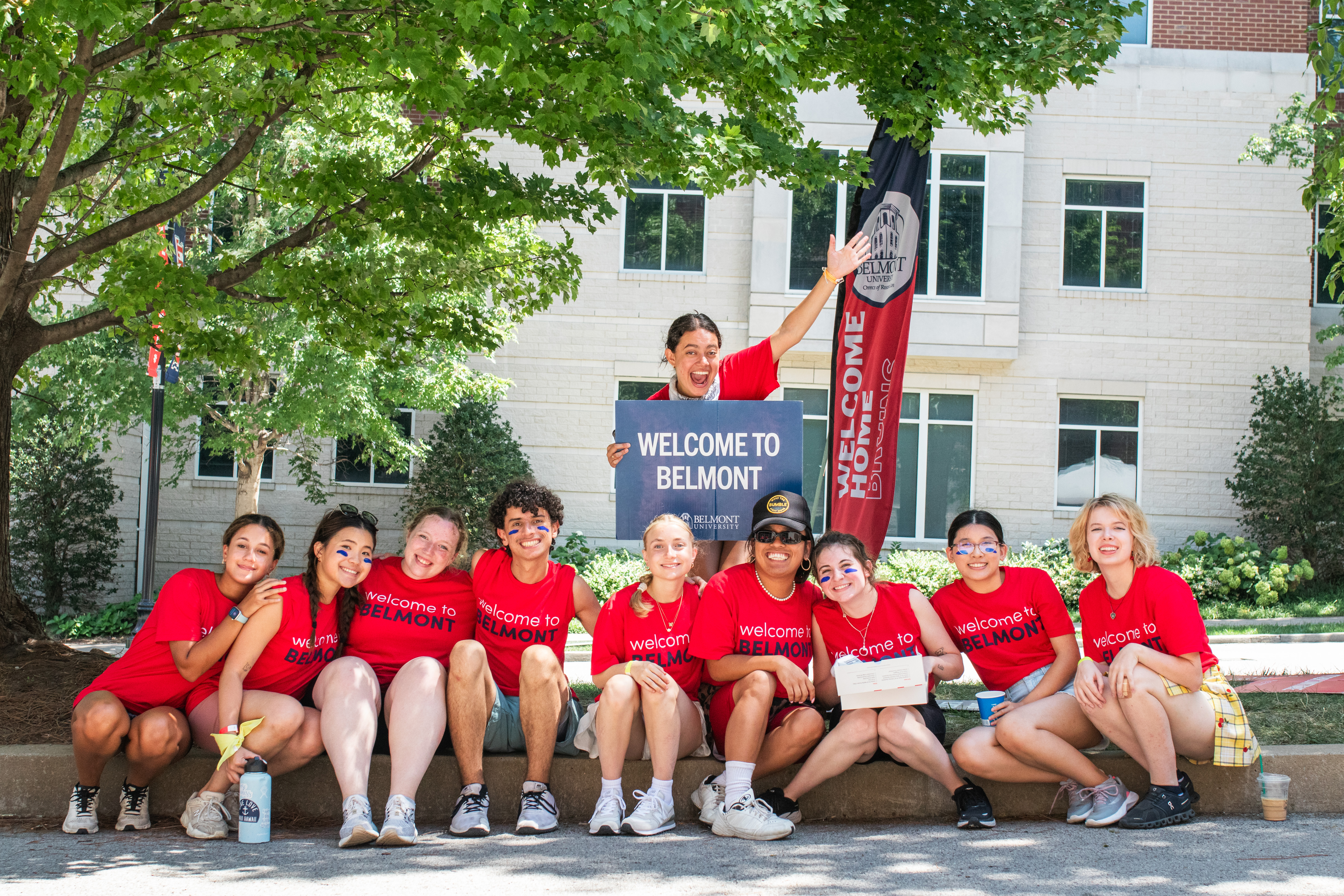 a group of ten students outside smiling wearing red welcome to belmont shirts and holding a welcome to belmont sign