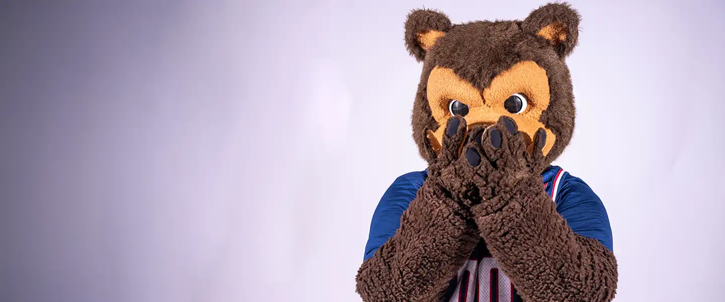 Belmont mascot Bruiser the Bruin covers his mouth with both paws while wearing a blue basketball jersey.