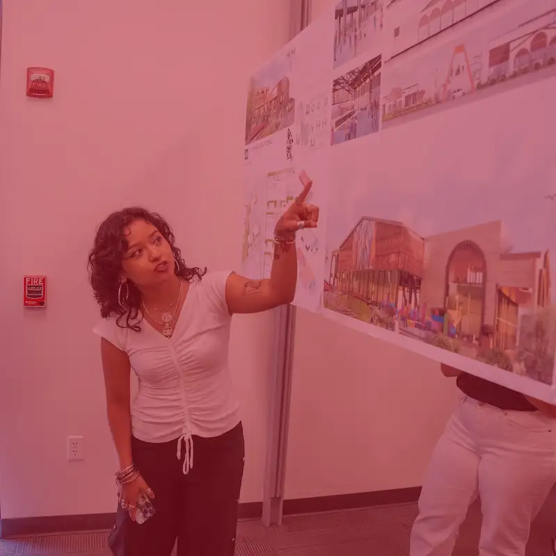 Student gestures while presenting architectural design board featuring colorful renderings and site plans.