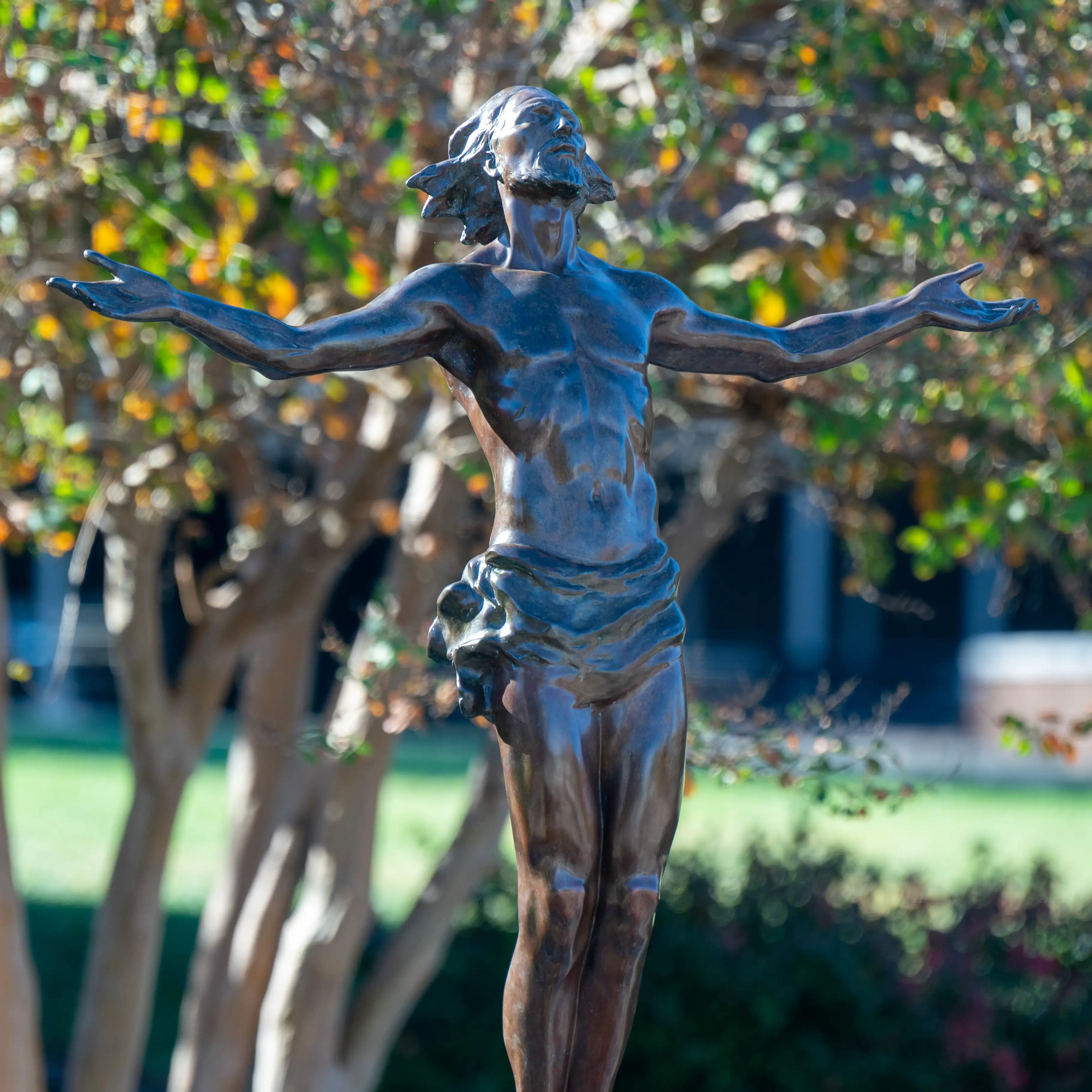 Bronze statue of Christ with outstretched arms on Belmont University’s campus, framed by trees and greenery.