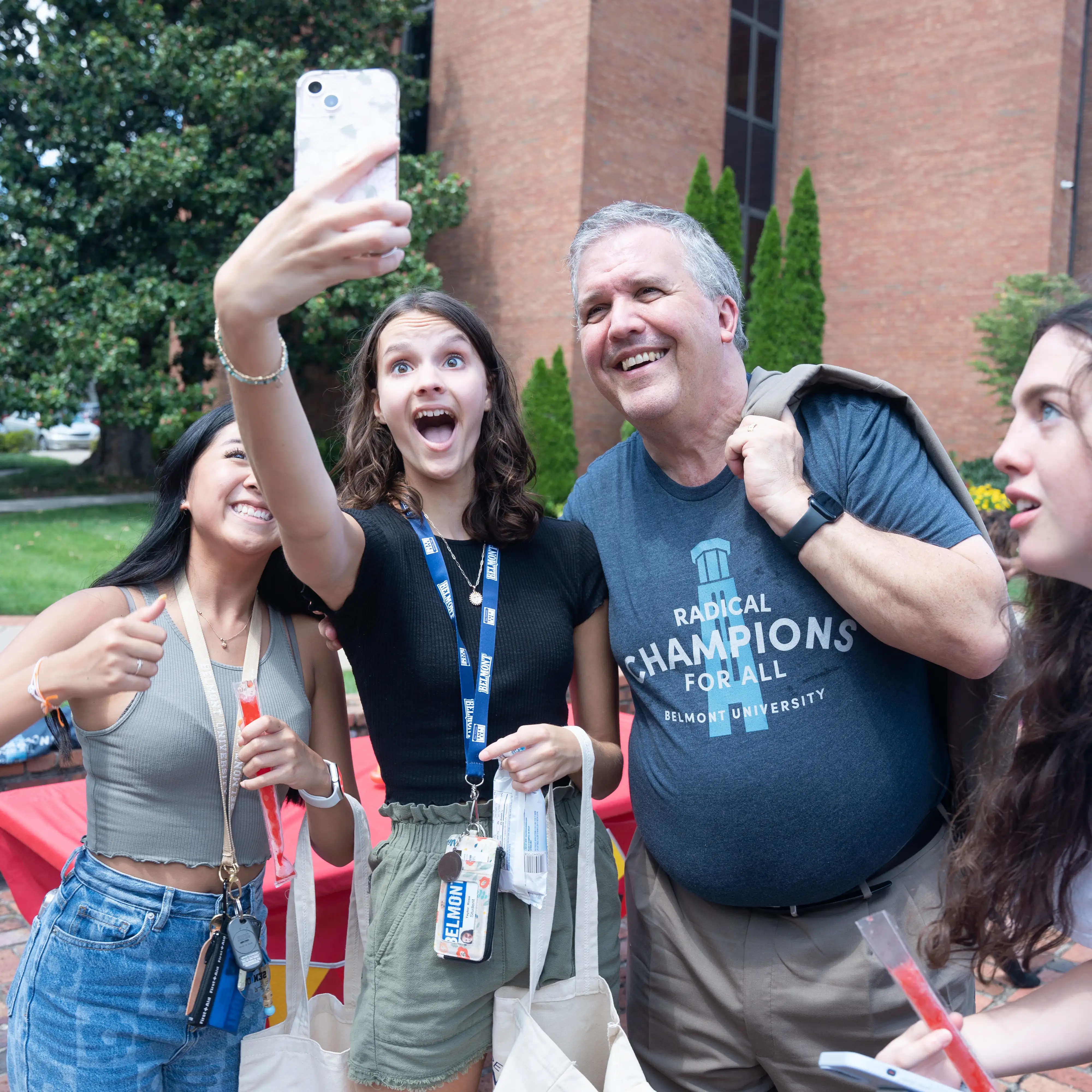 An outdoor photo of a group of female students and Greg Jones by the Bell Tower taking a group selfie with one girl holding her phone out in front of them.