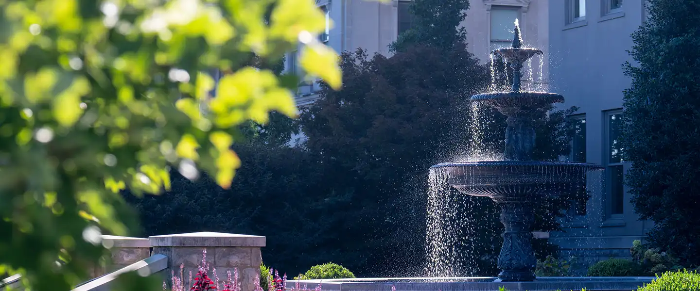 A decorative campus fountain surrounded by landscaping, with sunlight reflecting off cascading water and greenery in the background.