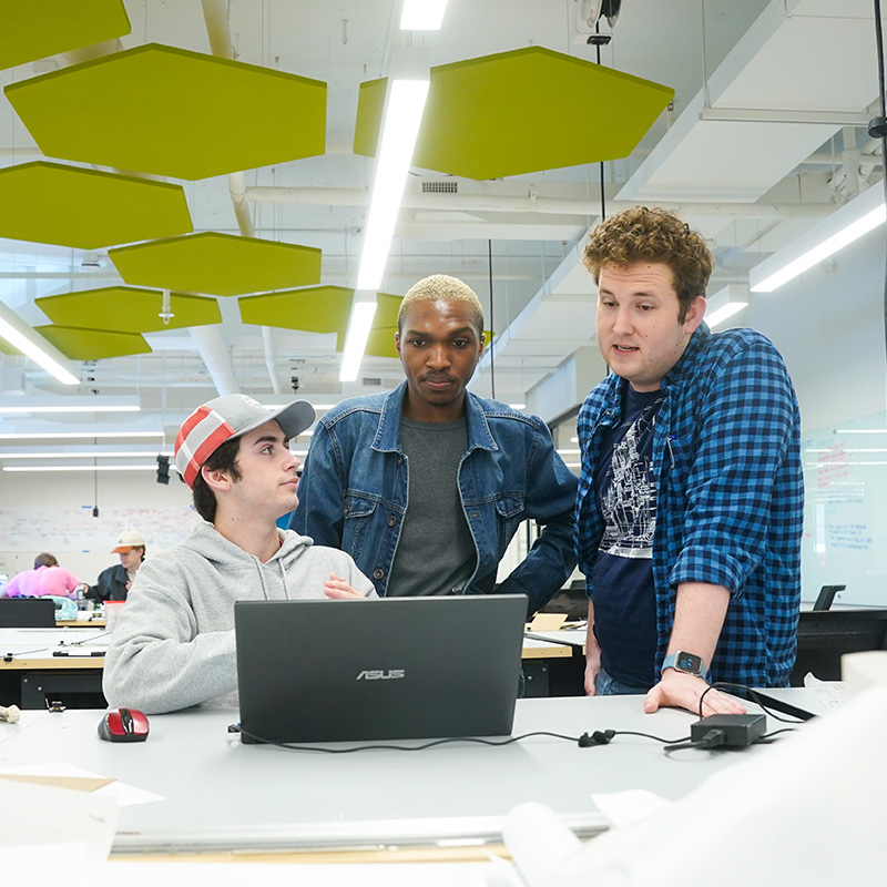 Two male students look over another student's shoulder as they work together from a laptop.