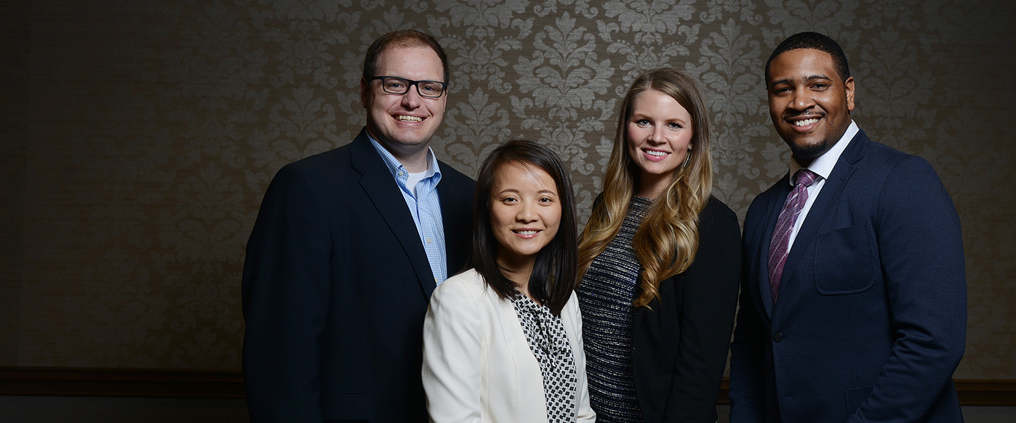 Four adult students dressed professionally stand together and smile at the camera.