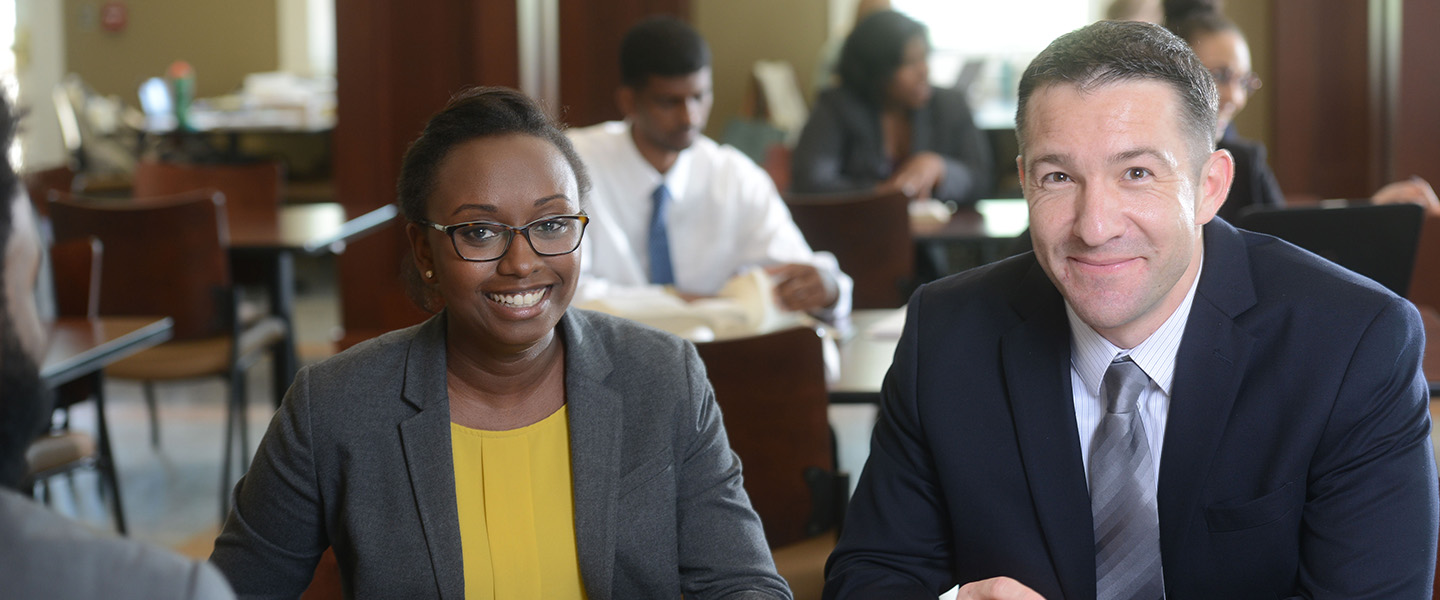 Two students dressed in suits sit in a wood-paneled room and smile.