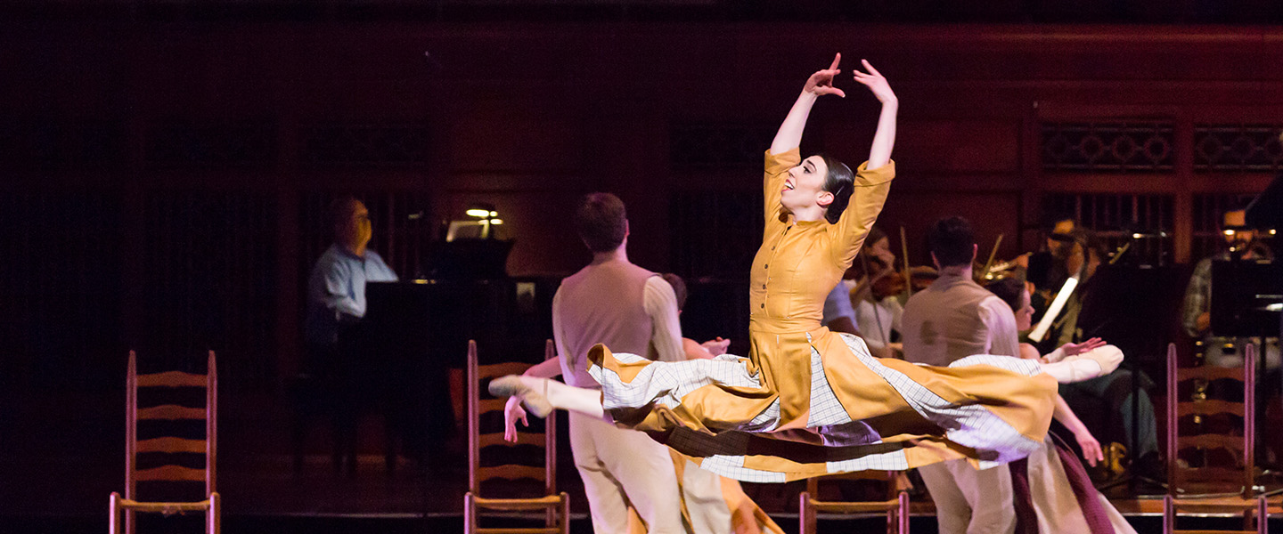 A female ballerina leaps through the air while dancing on stage.