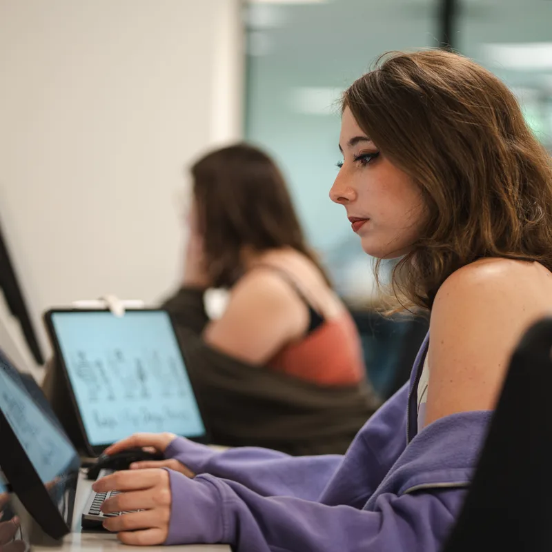 Young woman in profile working on a laptop in a classroom or office, with another student blurred in the background.