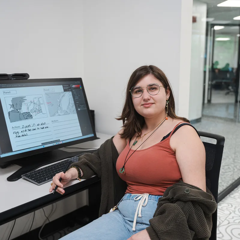 A woman with glasses sitting at a desk, working on a storyboard on a tablet displaying sketches and handwritten notes.