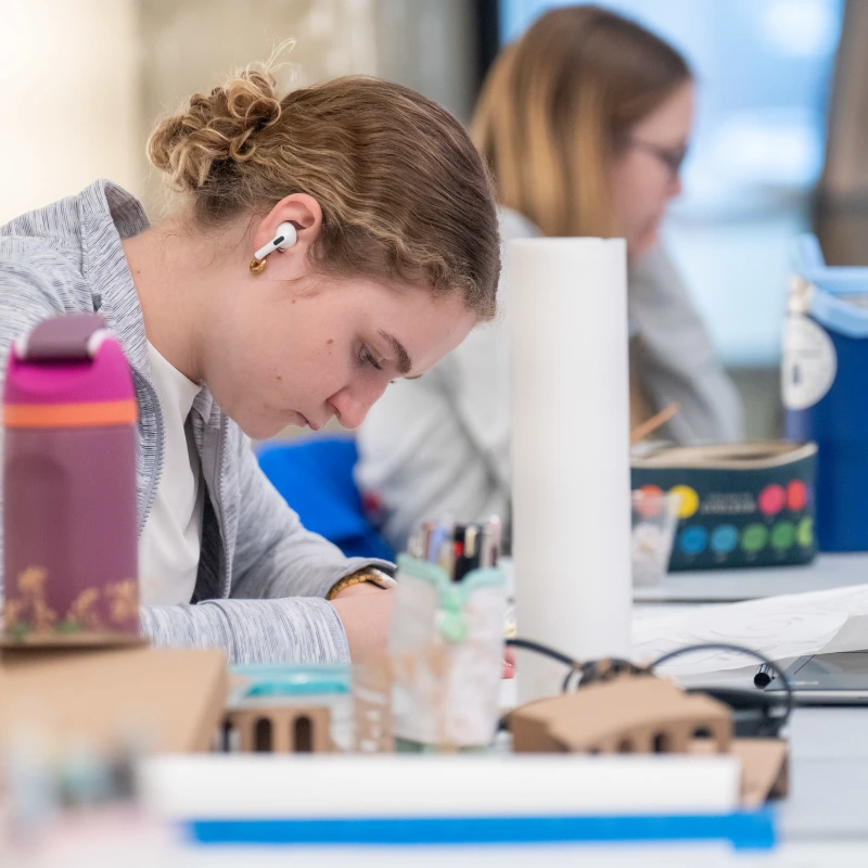 An architecture classroom scene featuring two students focused on their laptops, with design tools and sketches nearby.