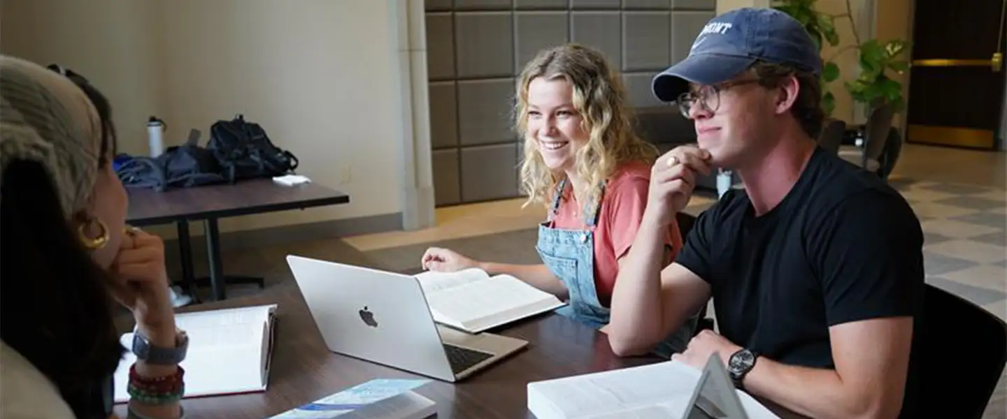 A group of three students engaged in a study session at a table, with laptops and books open. Two students are smiling, while one is listening intently.