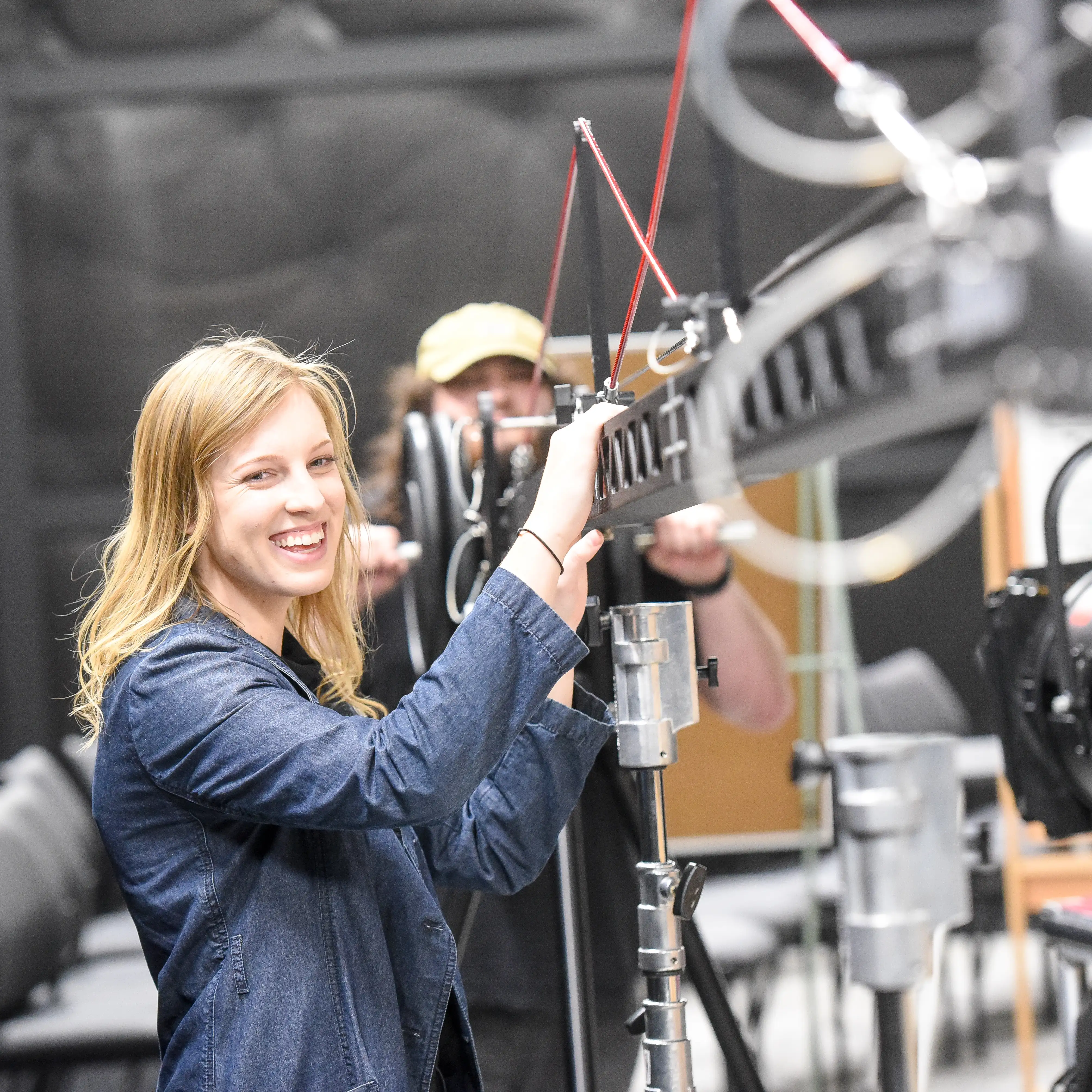 Smiling student adjusts a camera crane arm in a film production studio, with another student in the background assisting with equipment setup.
