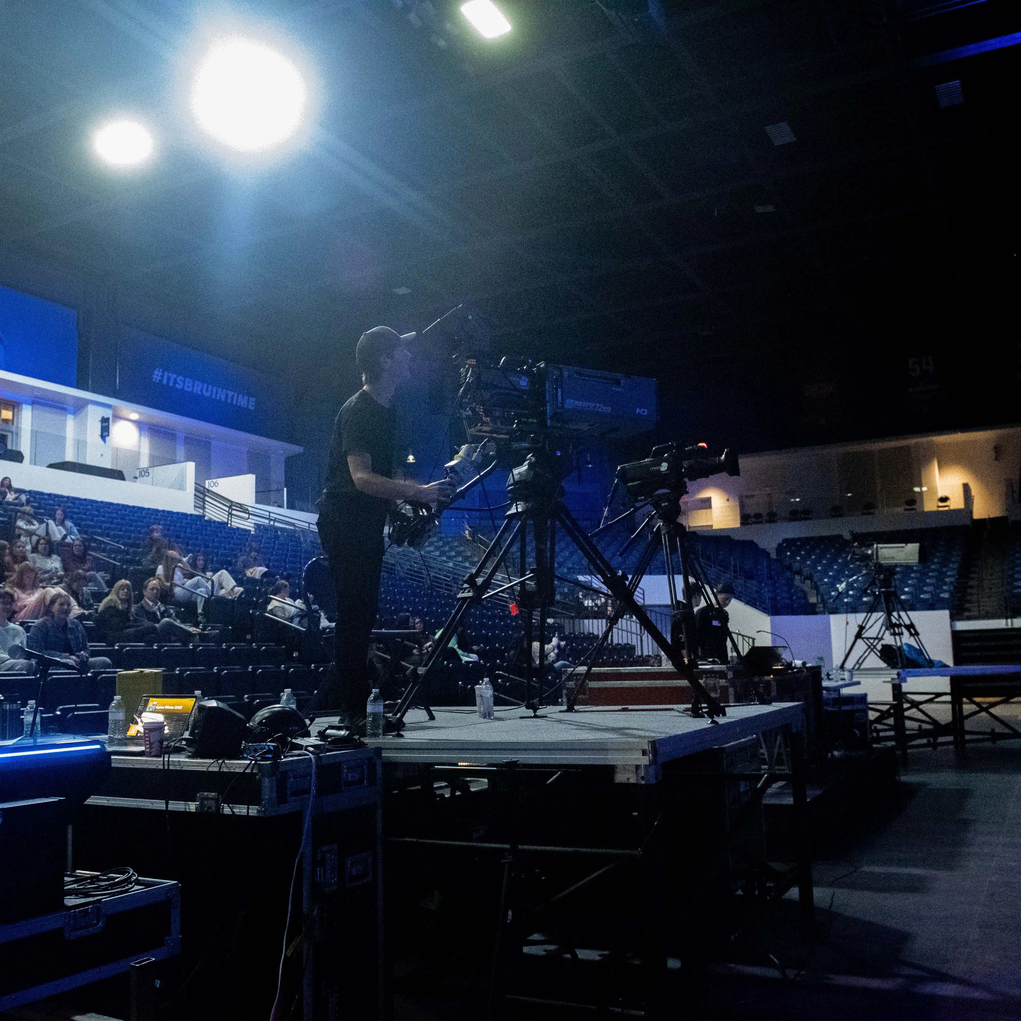 Camera operator films from a raised platform inside a large arena, with multiple cameras and audience members visible in the background.