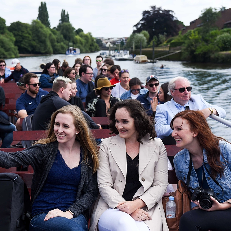 Three women smile and lean in together while sitting on an open-air riverboat filled with passengers. The woman on the right holds a camera, and lush green trees line the riverbanks in the background under an overcast sky.