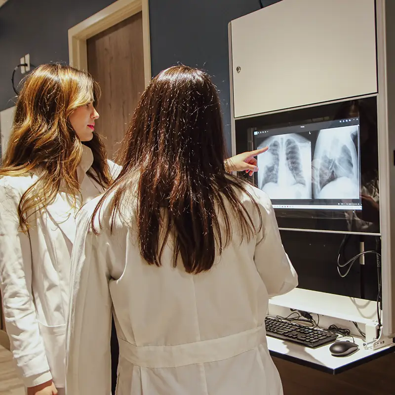 Two students in white lab coats examine X-ray images on a digital screen, with one pointing at the display while the other observes in a healthcare lab setting.