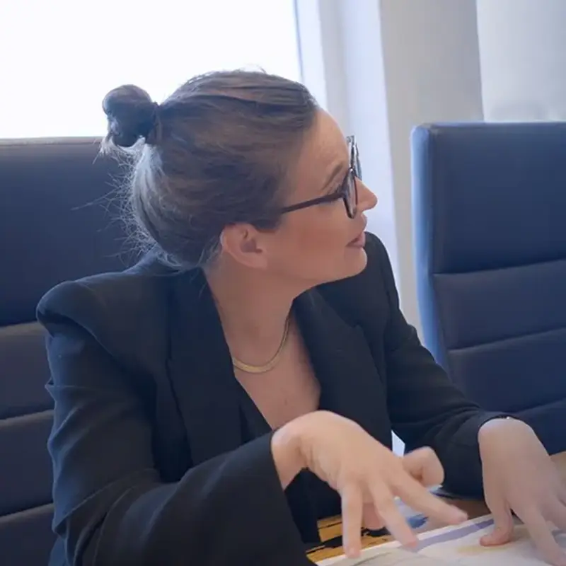 A woman in a business suit sits at a conference table, talking and gesturing with her hands while engaging in a conversation.