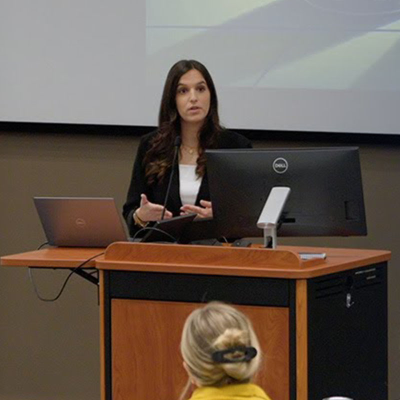 A student stands at a podium, speaking to a class while gesturing with her hands. A computer screen is visible on the podium.