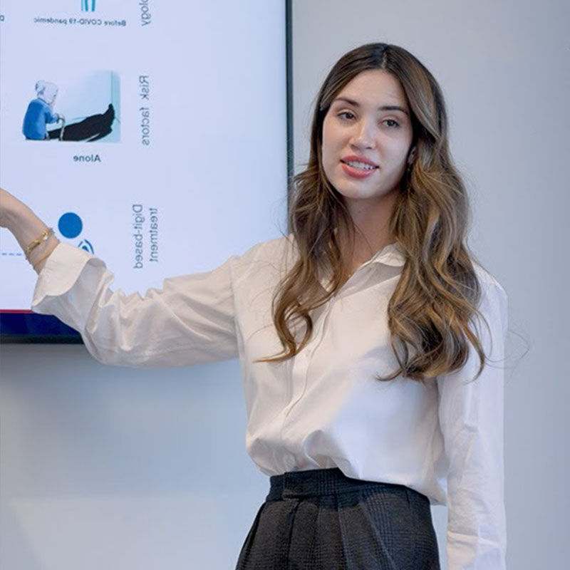 A student in a white blouse points to a presentation screen while explaining, with a clear focus on the visuals shown behind her.