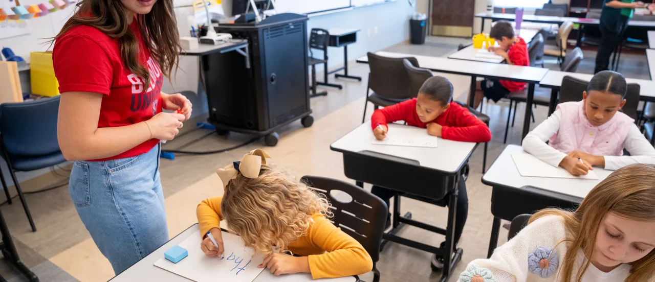 A young student sits next to a teacher in a field looking at a math workbook