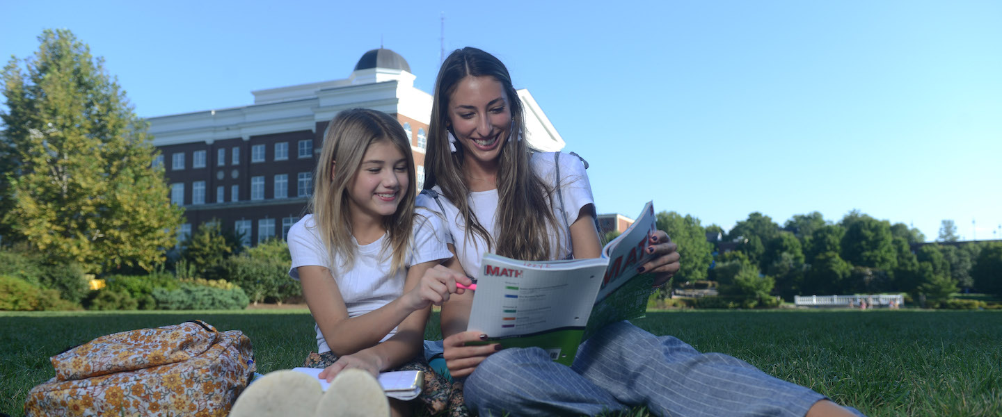 A young student sits next to a teacher in a field looking at a math workbook