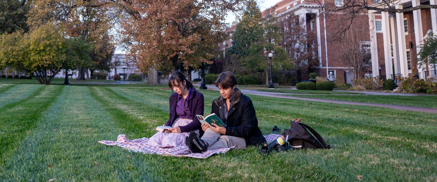 Two students read books on a vibrant green university campus lawn with autumn trees and a brick building.