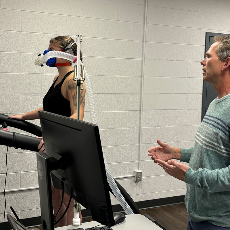 A woman running on a treadmill while wearing a breathing mask connected to a metabolic testing system, as a male instructor in a striped sweater observes and provides guidance.