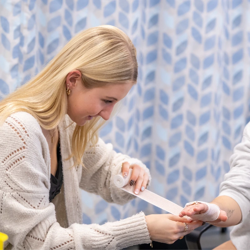 A woman with blonde hair wraps a bandage around another woman's hand. Both are seated, with a patterned curtain in the background.