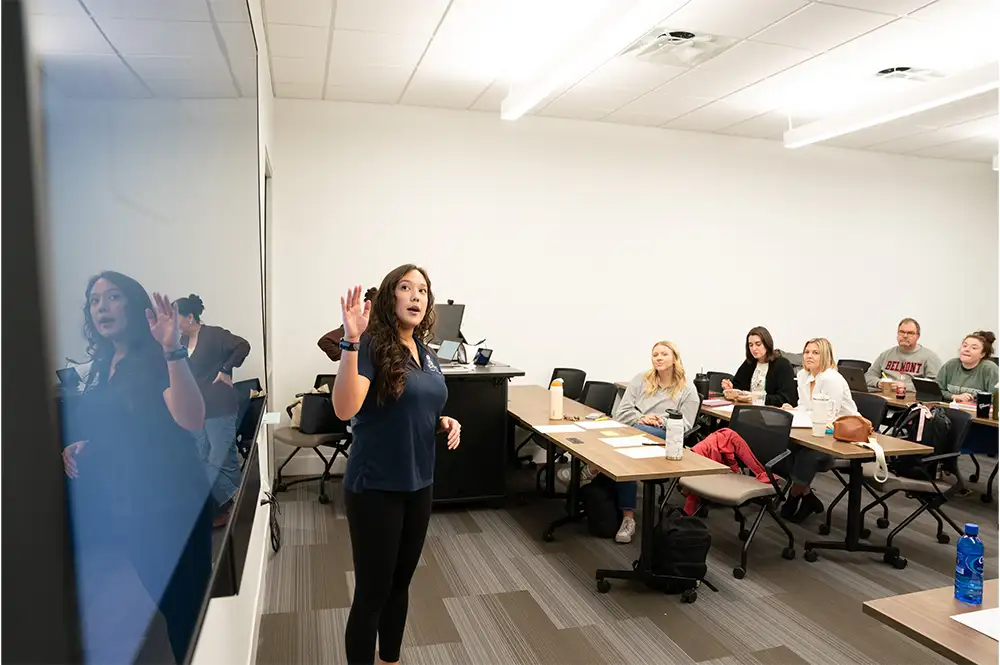 A classroom scene featuring a woman standing in front of a presentation screen, gesturing as she speaks to a group of attentive students seated at desks. The students, mostly women, are engaged and taking notes, with various personal items like water bottles and notebooks visible on their desks. The room has a modern feel, with bright lighting and a clean layout, enhancing the learning environment.