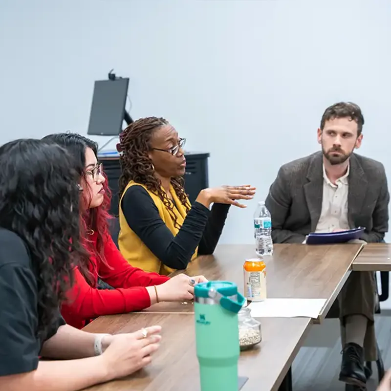 A group of people engaged in a discussion at a meeting table. Four individuals are seated, with one person actively speaking. The speaker has long, curly hair and is wearing a yellow top. Others include a person in a black shirt, another in a red top, and a man in a suit listening intently.