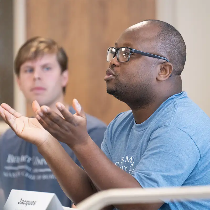 A student with glasses passionately engages in a discussion during a class session, gesturing with his hands to emphasize his points. In the background, another student listens attentively. The classroom setting reflects an interactive learning environment in the MBA and MACC programs.