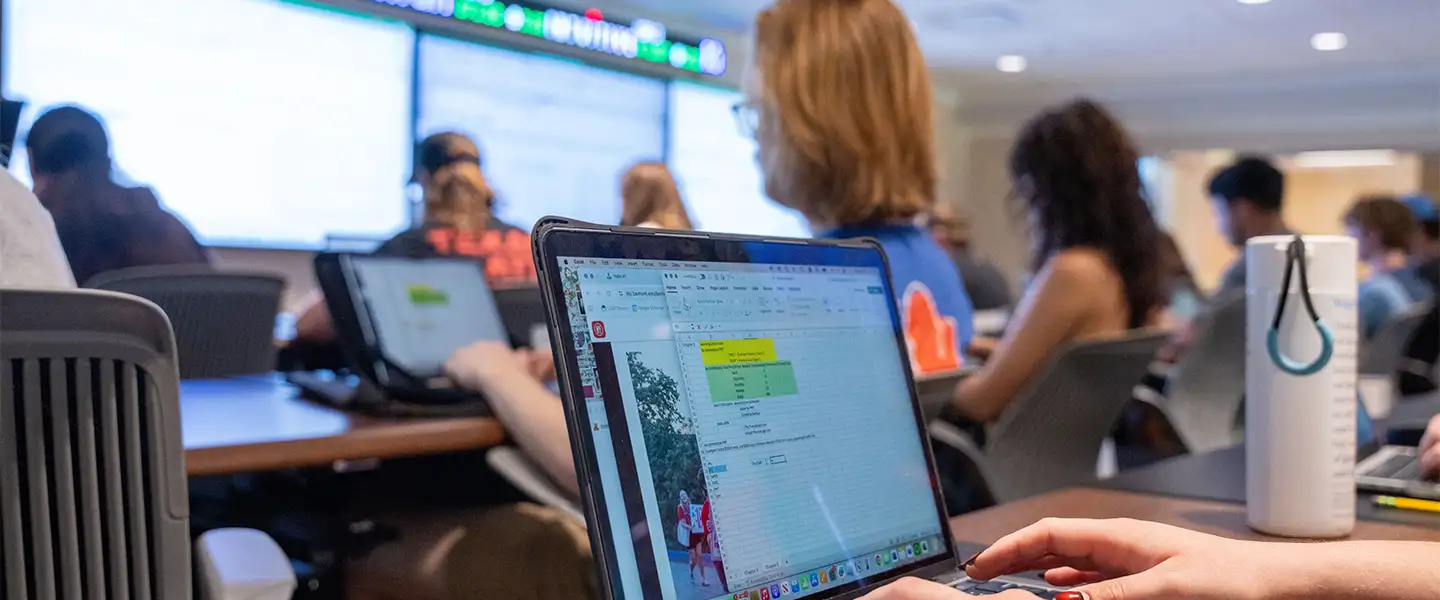 Students are engaged in a classroom setting, focusing on their laptops during a lecture. One student is seen typing, with a spreadsheet displayed on the screen, while large monitors in the background show relevant data. The atmosphere is collaborative and academic, highlighting the learning experience in the MBA and MACC programs.