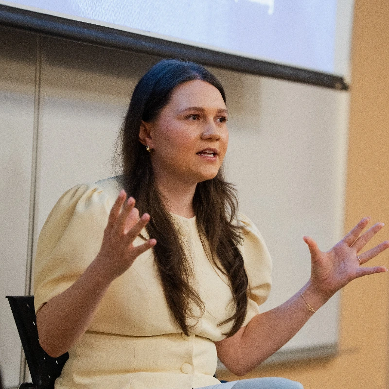 Woman speaking and gesturing animatedly during a presentation, wearing a light yellow top.