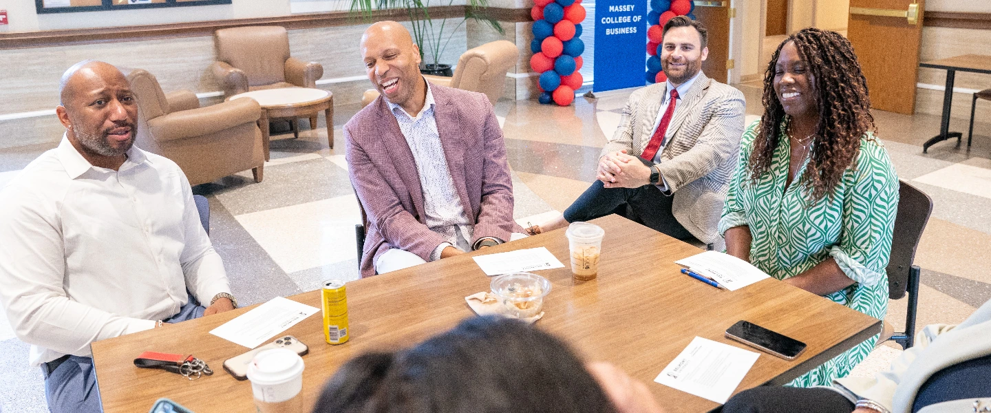 Four diverse professionals smiling during a meeting at a wooden table in a college of business.
