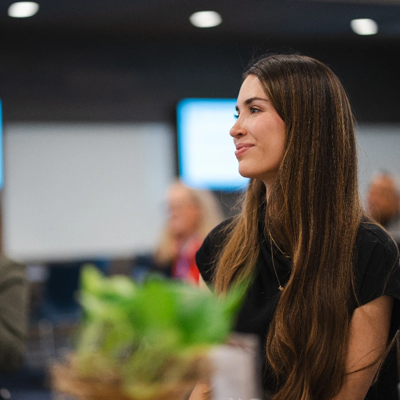 Smiling woman with long brown hair listening attentively at a professional conference event.