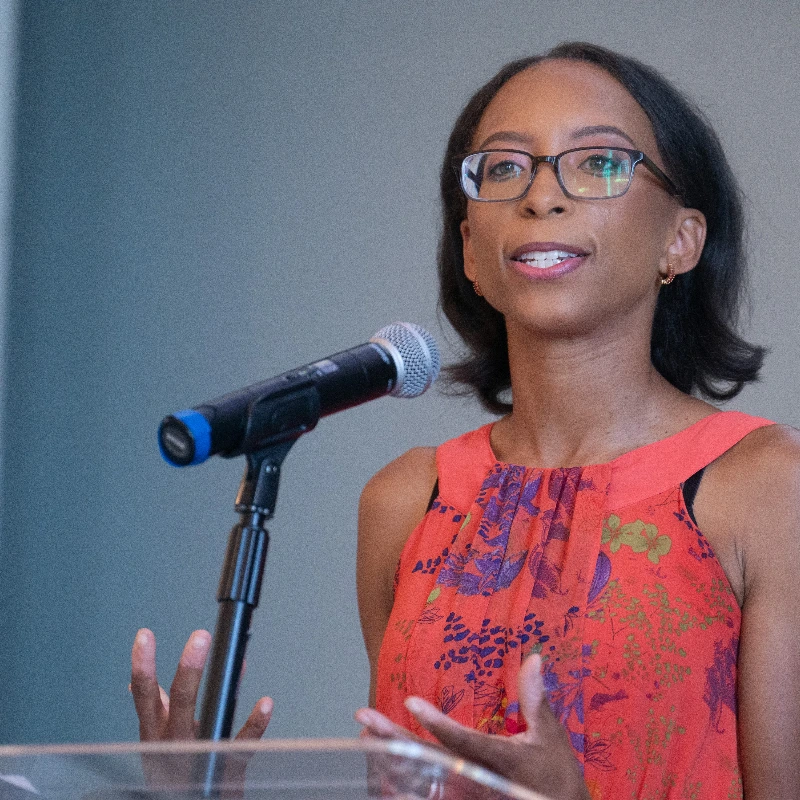 Woman with glasses speaking into a microphone at a podium during a presentation or conference.
