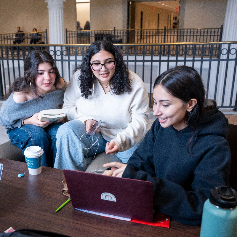 Three young women collaborating on a laptop in a modern university library, focused on their study project