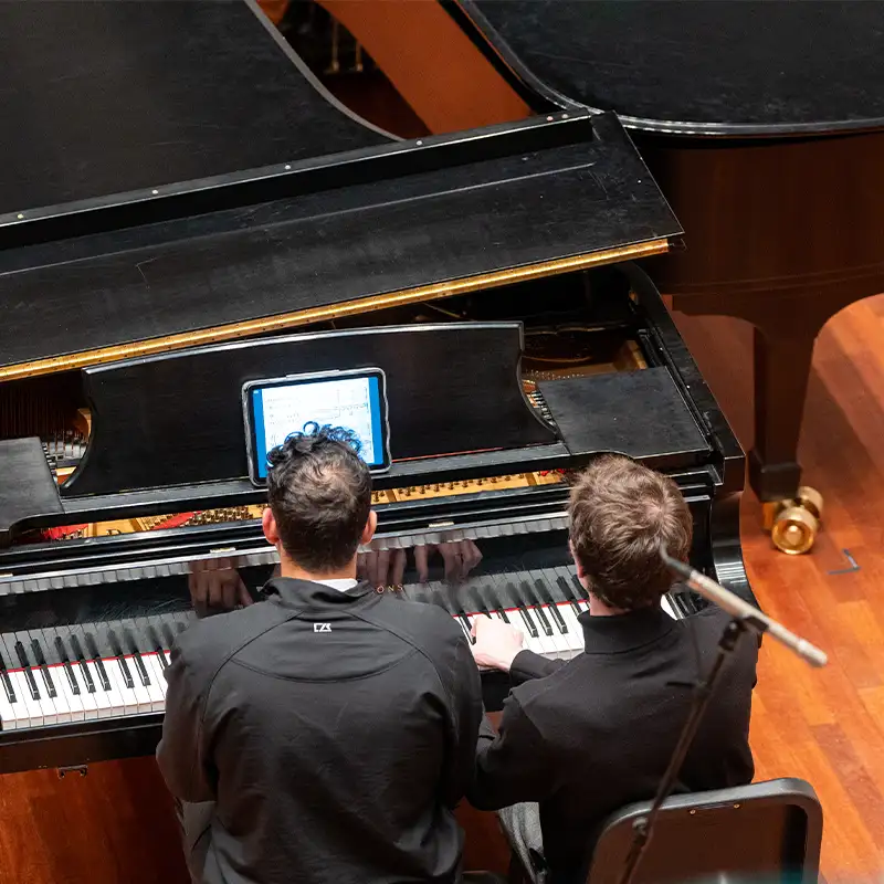 Two students in a keyboard pedagogy class collaborating at a grand piano, with one student playing and the other following along using a tablet placed on the piano.