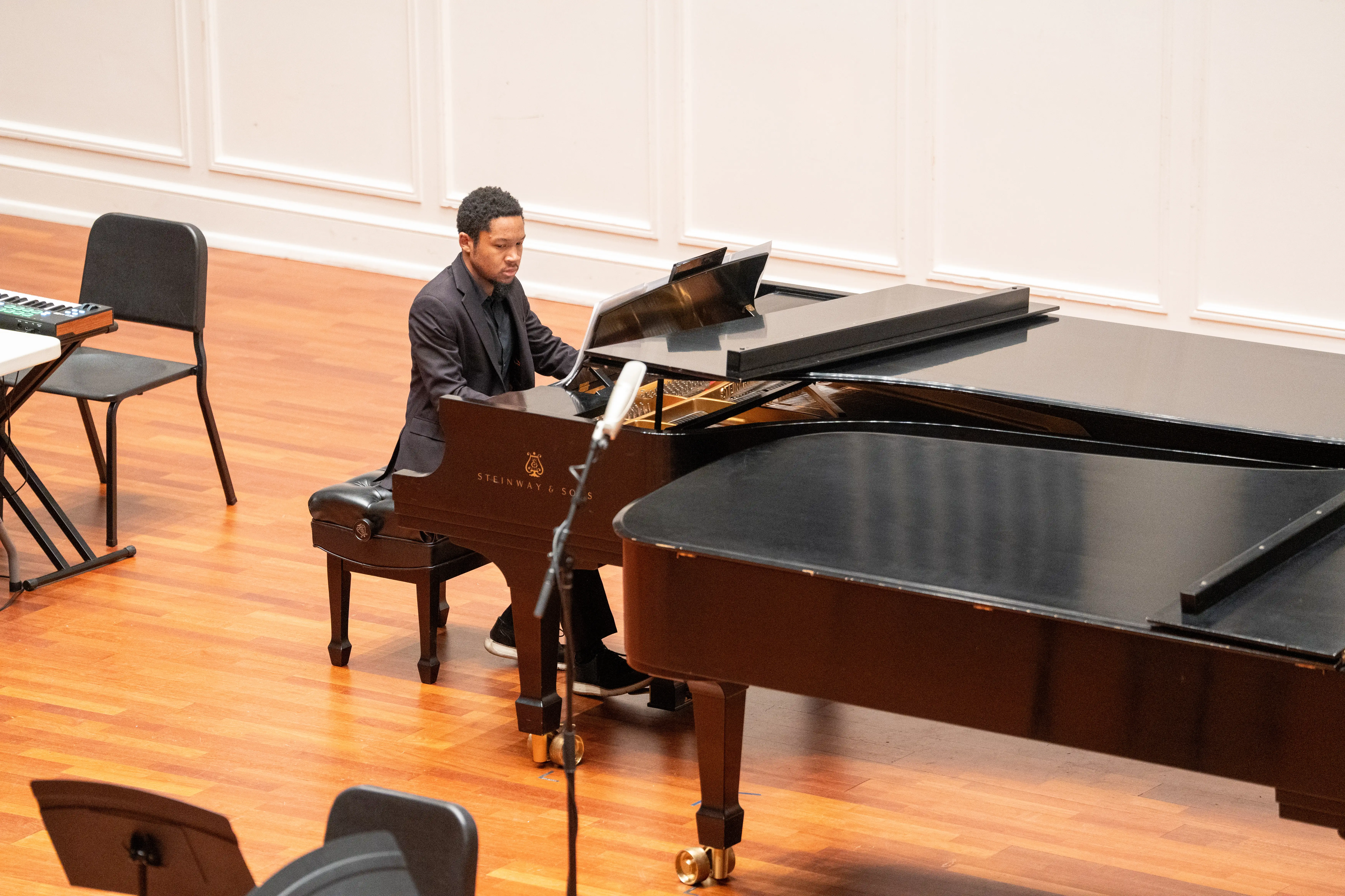 A student performing at a grand piano during a keyboard pedagogy class, dressed in formal attire, with a keyboard and empty chairs visible in the background.