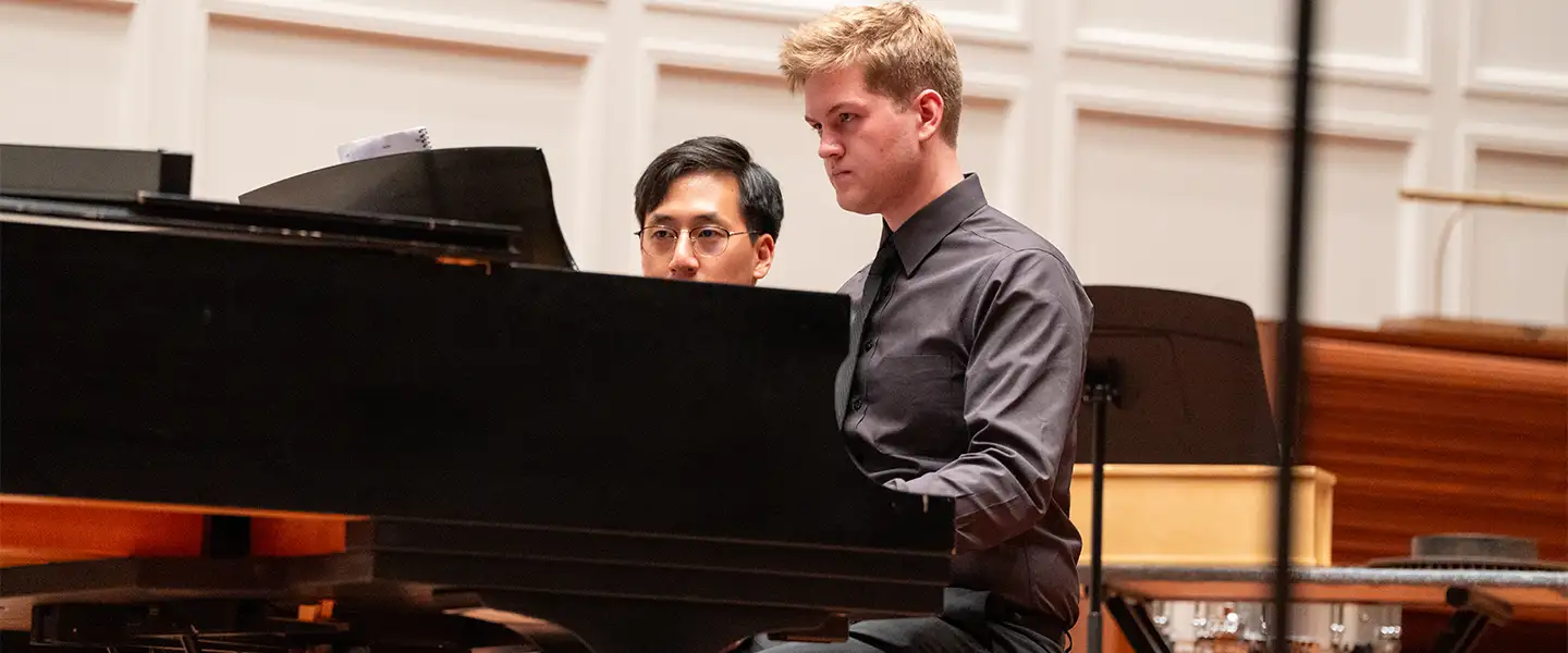 Two students in a keyboard performance class, collaborating at a grand piano. One student is playing while the other observes, both dressed in formal attire.