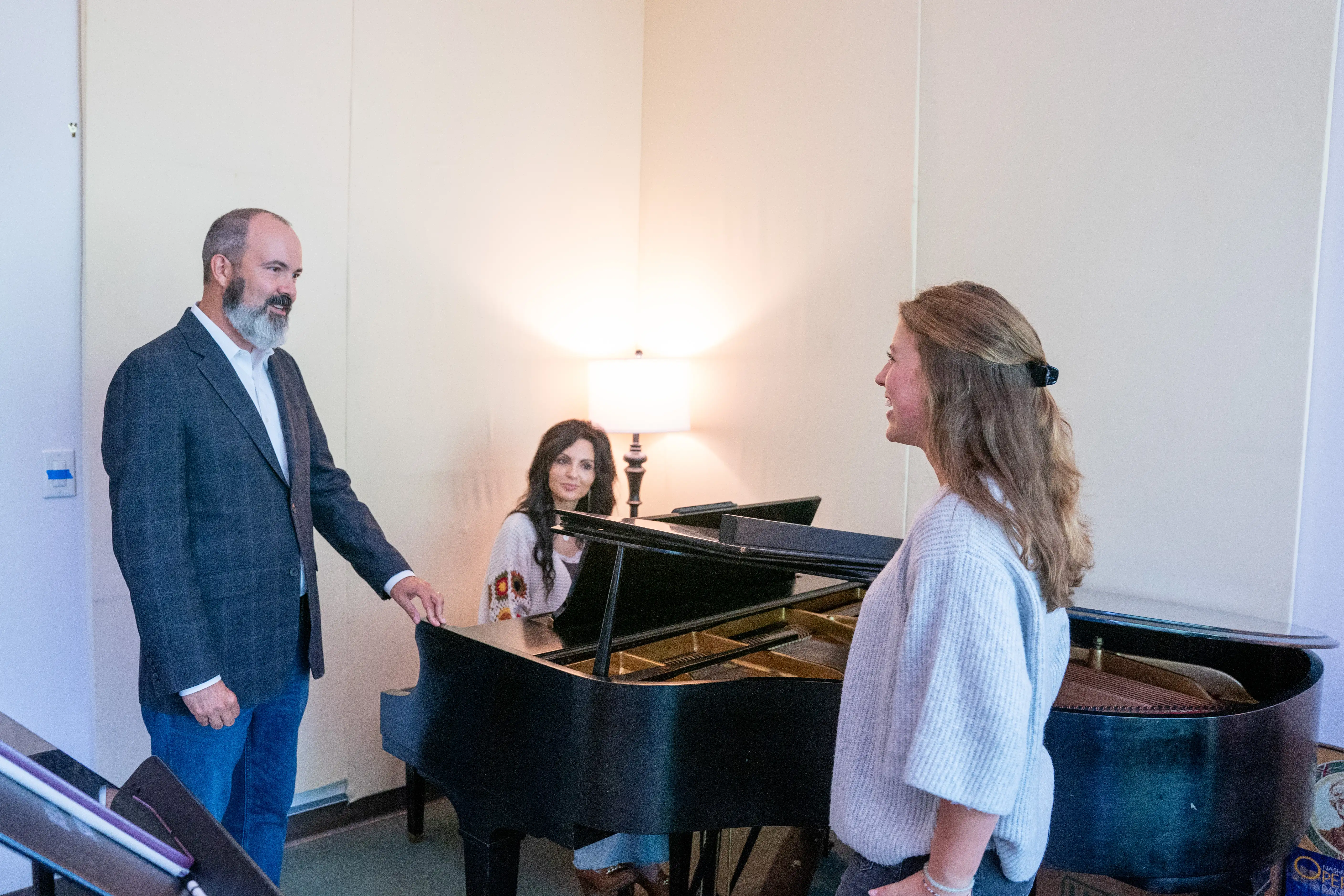A bearded vocal coach in a blazer gestures toward a grand piano while a pianist sits at the keyboard and a young woman stands singing beside the instrument in a bright practice room.