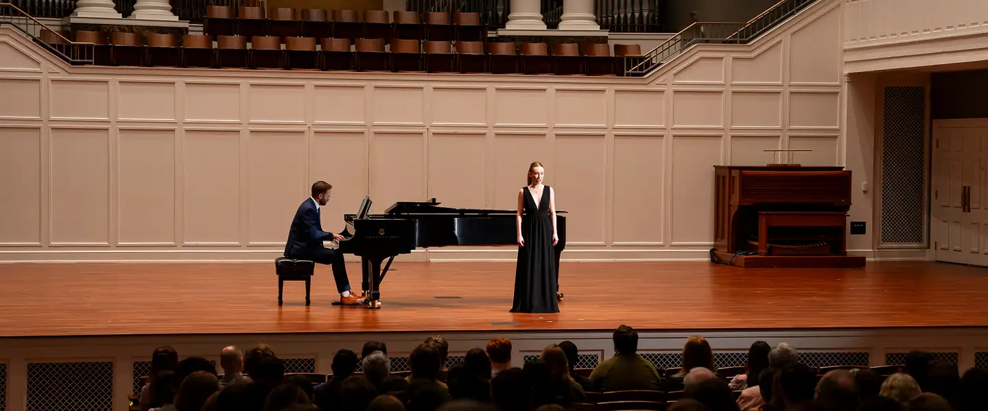 Pianist seated at a grand piano accompanies a soprano vocalist standing center-stage in McAfee concert hall, with audience silhouettes in the foreground.
