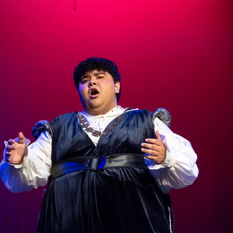 A male vocalist performing on stage in a dramatic pose, wearing a blue velvet costume with silver accents, against a vibrant pink background.