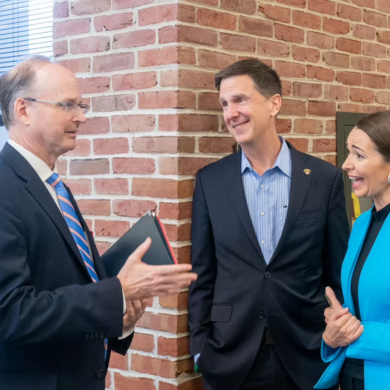 Three people in business attire are conversing cheerfully by a brick wall in an office setting. One man gestures while others smile and listen.