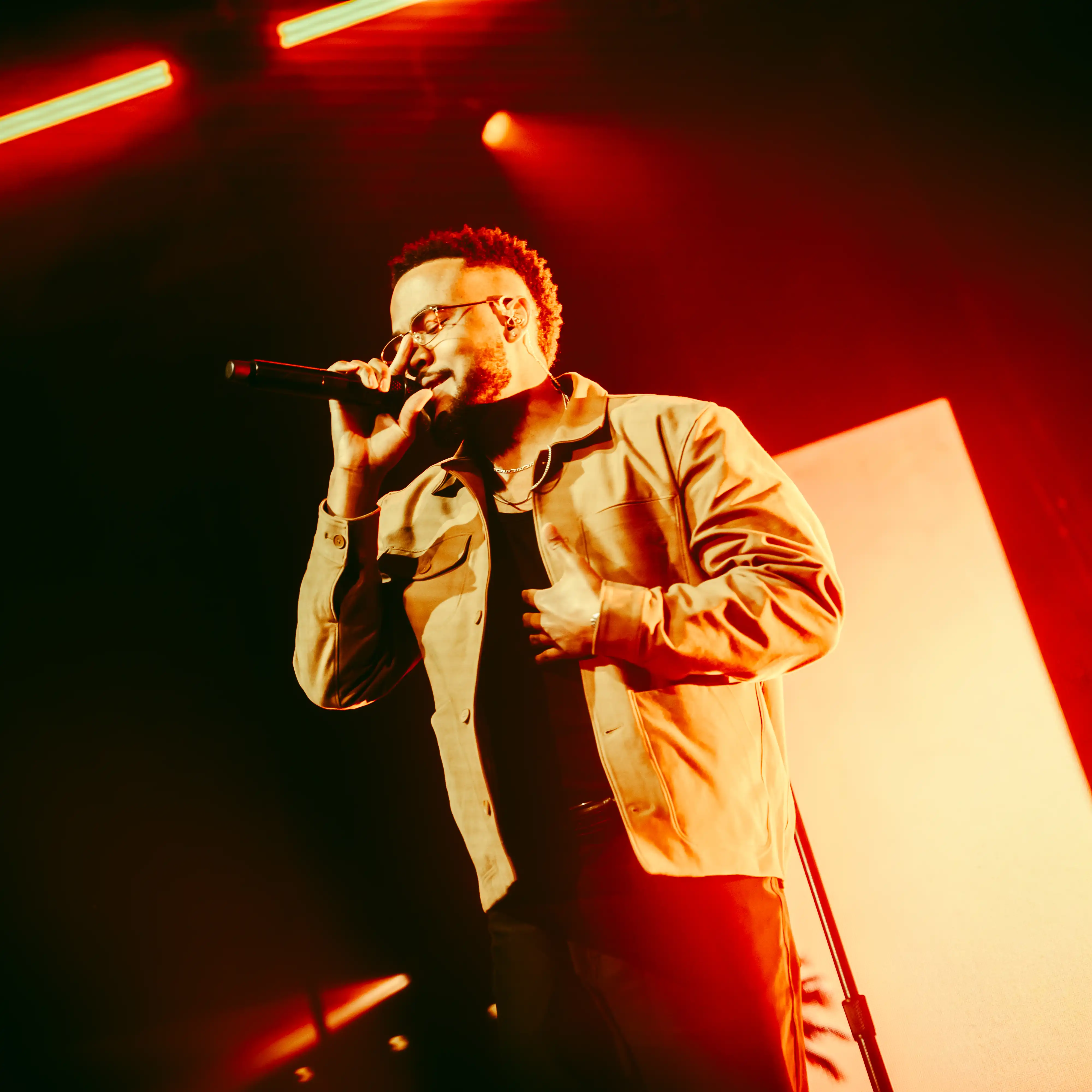 Performer sings into a microphone onstage under dramatic red lighting during a live music performance.