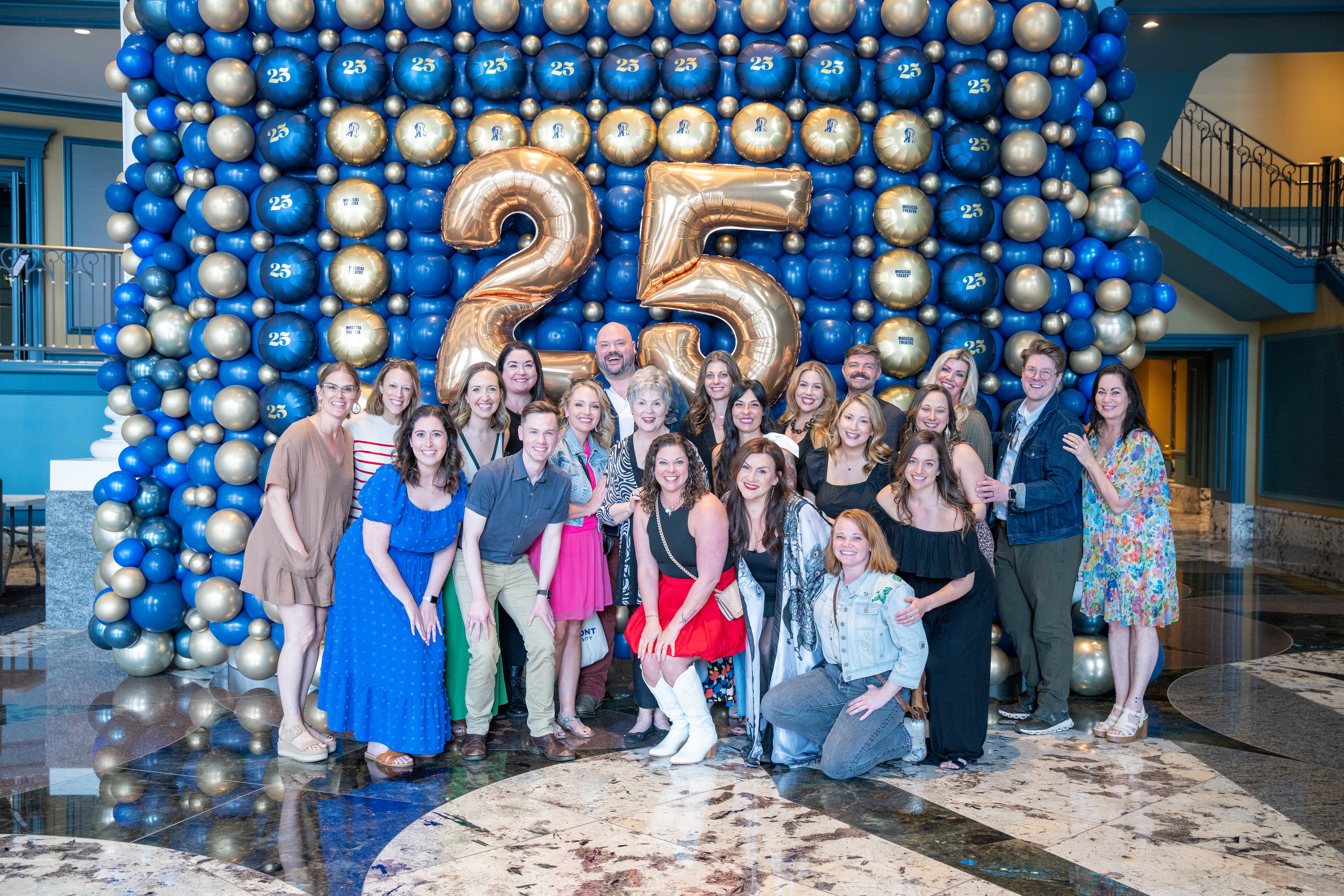 25th Anniversary Luncheon for Musical Theater, 22 people posed in front of blue and gold balloons 