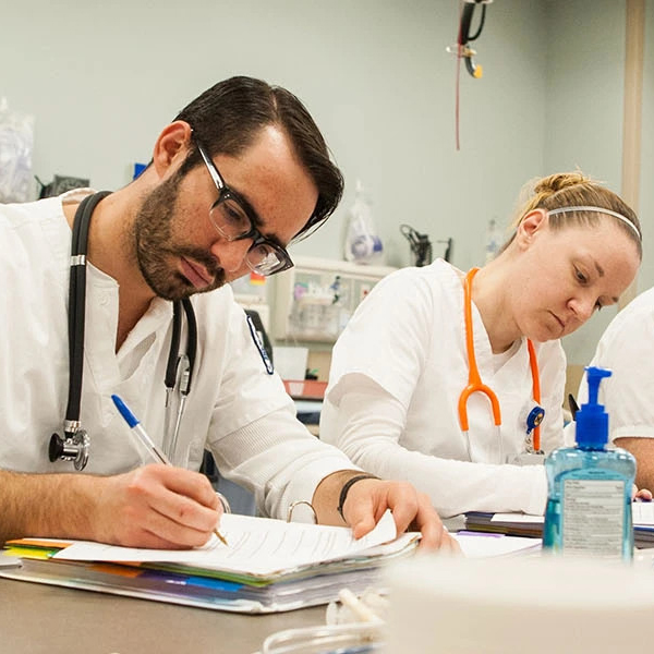 Students in a nursing classroom setting write notes at a table, wearing white clinical tops and stethoscopes as part of academic coursework.